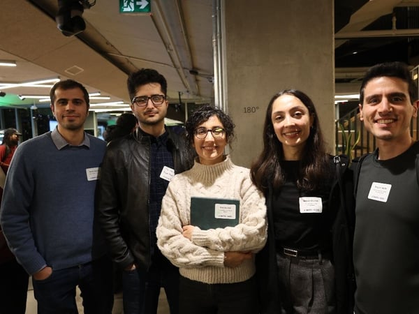 A group of five people, consisting of three men and two women, stand together and smile directly at the camera in what appears to be an indoor professional or networking event.