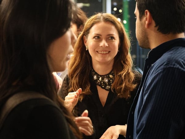 A close-up, candid photograph captures a smiling woman with long brown hair and a large jeweled necklace engaged in conversation with two other people in a dimly lit indoor setting.