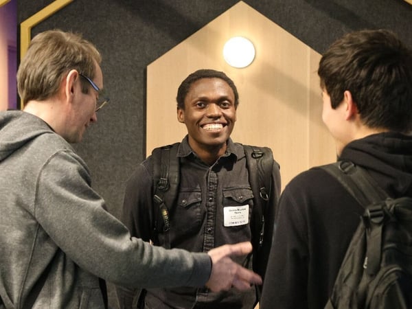 A candid indoor photograph captures a young man smiling widely while interacting with two other individuals, possibly during a professional or networking event. The man in the center wears a name tag and stands against a modern backdrop featuring geometric wood and felt panels.