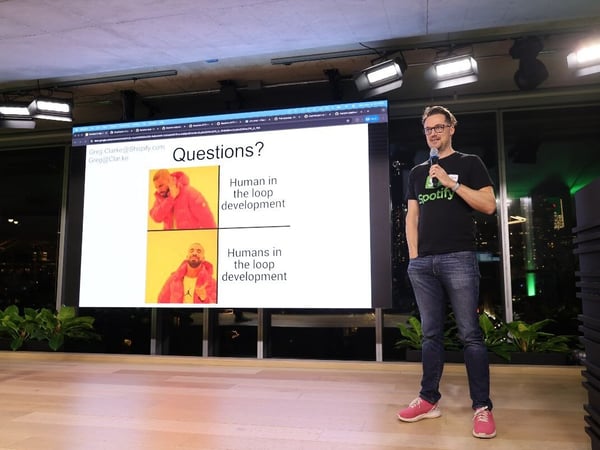 A man wearing a Spotify t-shirt and glasses is giving a presentation on a stage, holding a microphone, with a large screen behind him displaying a slide about 'Human in the loop development' using the Drake meme format.