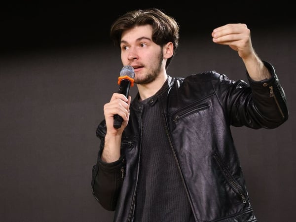 A young man wearing a black leather jacket and a dark ribbed sweater is speaking into a microphone while gesturing emphatically with his left hand against a dark, featureless background.