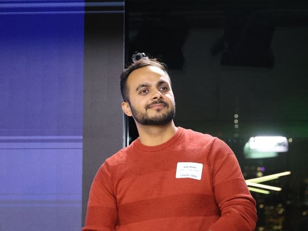 A candid photo captures a man, Amit Parekh, wearing a reddish-orange striped sweater and a nametag, standing in a dimly lit indoor venue with a large blue screen visible on the left.