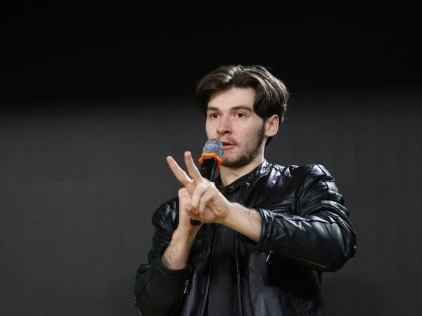 A young man wearing a black leather jacket is captured in a close-up shot while holding a microphone and making a peace sign gesture during a presentation or performance against a dark, featureless background.