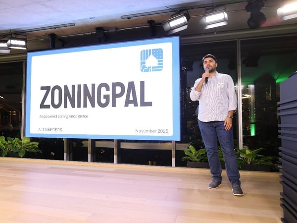 A man stands on a stage holding a microphone, giving a presentation in front of a large screen that displays the title slide for "ZONINGPAL."