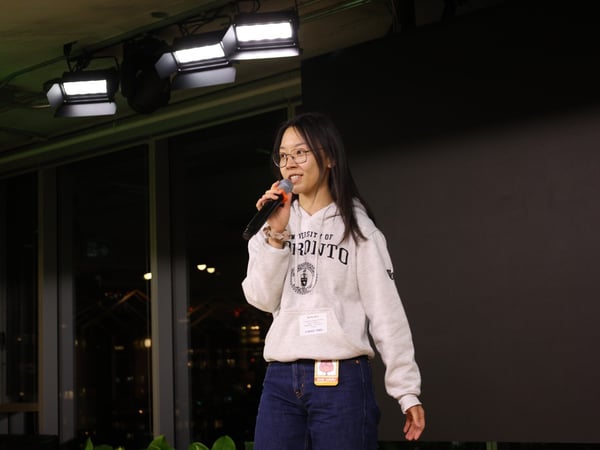 A young woman wearing a grey University of Toronto hoodie and glasses is speaking into a black microphone on a stage during an evening event. The background features stage lighting above, a dark screen to the right, and large windows revealing a city skyline at night.