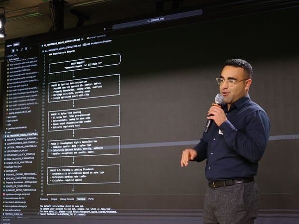 A photograph showing a man presenting technical information, speaking into a microphone while standing next to a large screen displaying an architecture diagram and code editor interface.