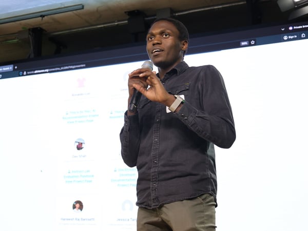 A young Black man, wearing a dark denim shirt, is giving a presentation or talk, holding a microphone while standing in front of a large projection screen displaying a web application interface.