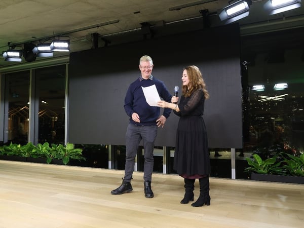 A photograph showing a man and a woman standing on a stage indoors at night, likely hosting an event or presentation. The woman holds a microphone, and the man holds a piece of paper, set against a large black screen and large windows overlooking a city skyline.