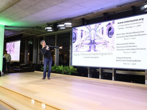 A man is giving a presentation on a wooden stage, speaking into a microphone and pointing toward a large projection screen that displays event information and a surreal image of a mirrored interior space.