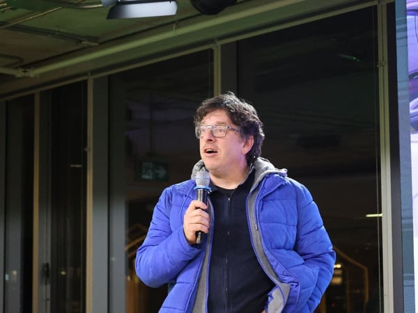 A photograph captures a man wearing glasses and a bright blue puffer jacket speaking into a microphone during an indoor event or presentation. He is standing in front of large dark windows or panels, with a green exit sign visible in the background.