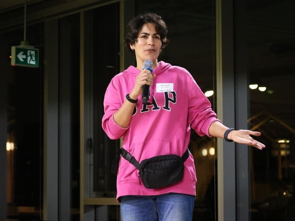 A woman wearing a bright pink GAP hoodie, blue jeans, and a black fanny pack is speaking into a microphone during an indoor event. She is standing in front of large glass panels, with the background appearing dark due to low lighting.