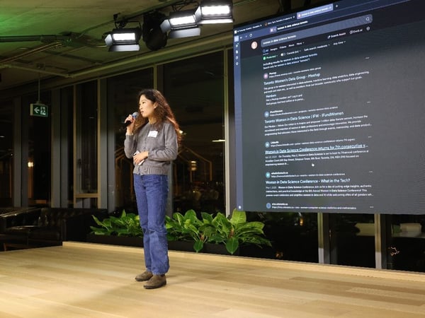 A woman stands on a wooden stage holding a microphone, giving a presentation in a dimly lit venue, with a massive digital screen behind her displaying Google search results about 'women in data science toronto.'