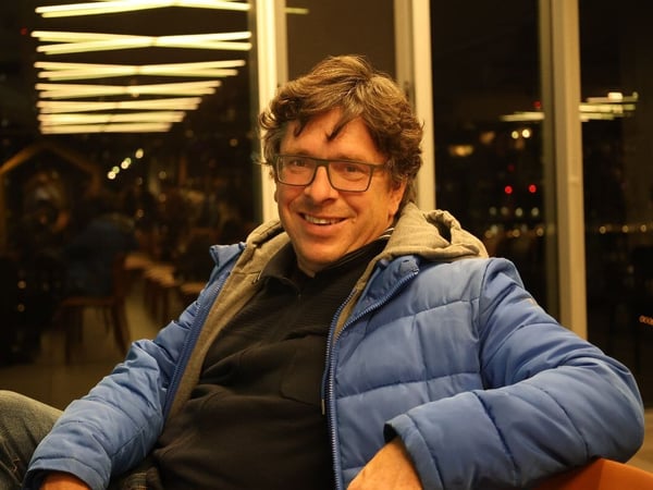A portrait photo captures a smiling middle-aged man wearing glasses and a blue puffer jacket, seated indoors at night against a dark background featuring blurred city lights and interior reflections.