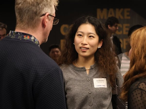 A candid photograph capturing a networking event where a young woman, Christina Zhou, is engaged in conversation with a man whose back is to the camera and another woman partially visible on the right, set against a dark background with large gold text signage.