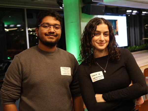 A portrait photo captures two young adults, a man and a woman, smiling directly at the camera while standing side-by-side at an indoor evening event characterized by green accent lighting.