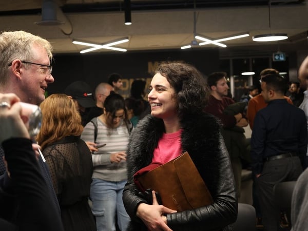 A candid photograph capturing a lively conversation between a man wearing glasses and a smiling woman in a black fur-trimmed jacket, holding a brown portfolio, at a crowded indoor social or networking event.