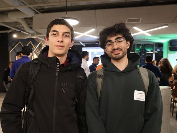 A candid indoor photograph captures two young men standing side-by-side, looking directly at the camera, likely at a professional or social event. The background shows other attendees mingling in a modern space with industrial ceilings and green accent lighting.