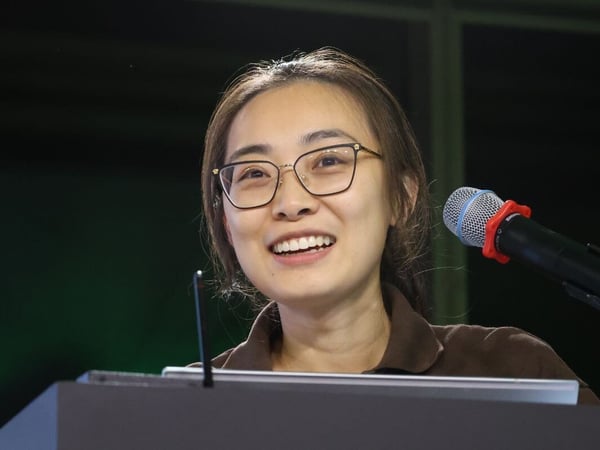 A close-up photograph captures a young woman with glasses, smiling widely, as she speaks into a microphone at a podium during an indoor presentation.