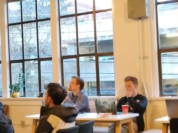 A candid photograph captures several people seated around a light wooden table in an indoor meeting or workspace, illuminated by large, multi-paned windows overlooking an urban setting. A mobile phone's volume and notification control overlay is visible on the right side of the image.