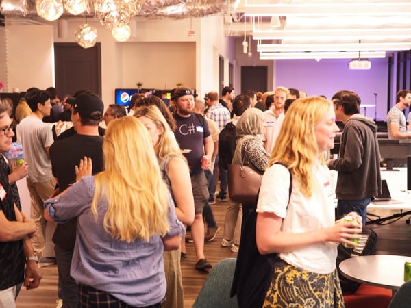 A candid photograph capturing a large crowd of people socializing and networking during an indoor event, likely a tech meetup, held within a modern office or event space.