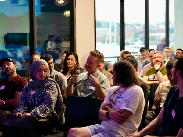 A photograph captures a diverse audience attending an indoor event, where a man in the center is holding a microphone and speaking or asking a question while gesturing with his hand. The room features large windows in the background overlooking an urban or waterfront area.