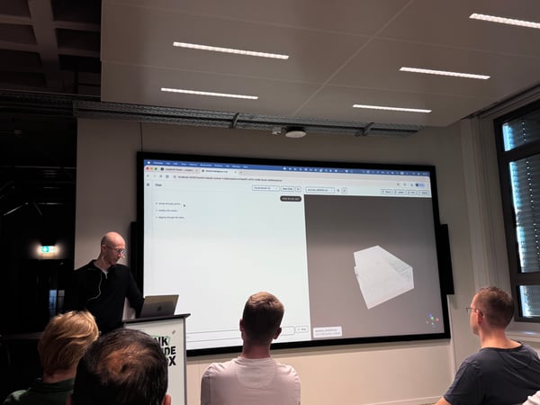 A man stands at a podium giving a presentation to an audience, with a large screen behind him displaying a 3D model and a chat interface.