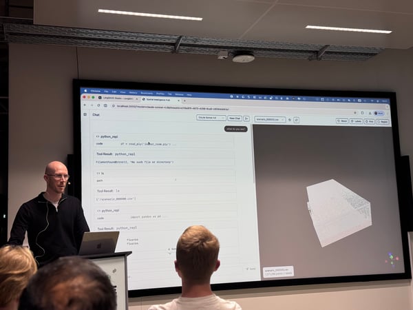 A man stands at a podium giving a presentation in front of a large screen displaying a coding interface and a 3D point cloud model.