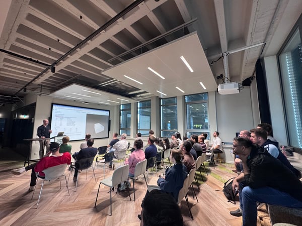A group of people are seated in a modern indoor space, attending a presentation where a man stands next to a large screen displaying technical data and a 3D model.