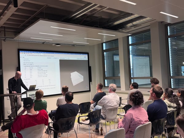 A man is giving a presentation to an audience in a modern office or classroom setting, with a large screen displaying data and a 3D model.