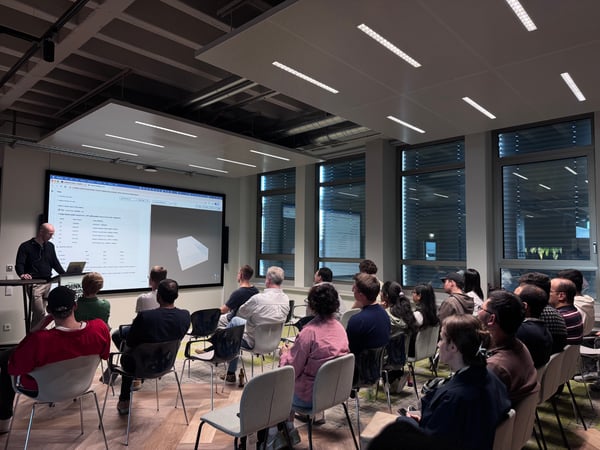 A man stands at a podium giving a technical presentation to a seated audience in a modern office or classroom environment.