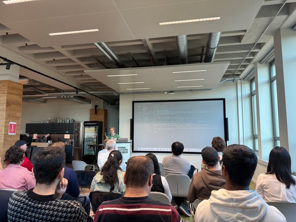 A group of people in a modern office space attending a technical presentation where code is displayed on a large screen.