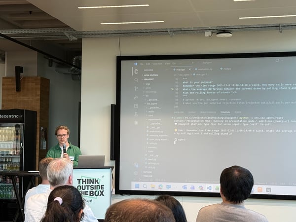 A man in a green shirt is giving a presentation in front of a large screen displaying code and a terminal window, while an audience watches from the foreground.
