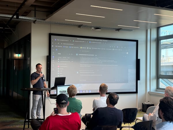 A man is giving a presentation in a modern office space, showcasing a software demo on a large screen to a seated audience.