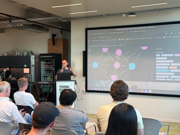 A man stands at a podium giving a technical presentation about graph databases to a seated audience in an office environment.