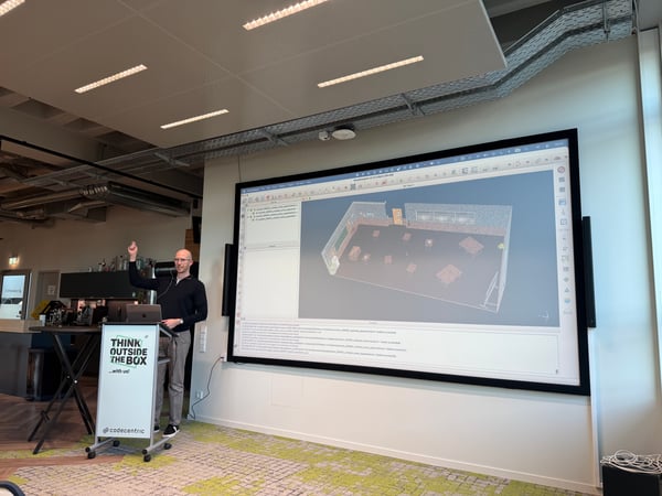 A man stands at a branded podium giving a presentation, with a large screen behind him displaying a 3D room model.