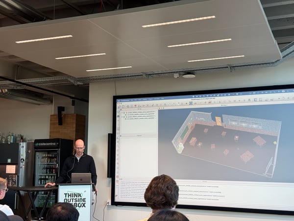 A man is giving a technical presentation in an office setting, standing behind a podium with a laptop in front of a large screen displaying a 3D room model.