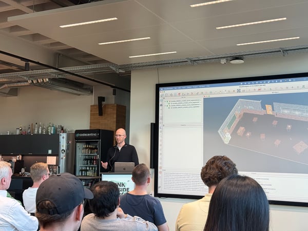 A man is giving a presentation in a modern office space, standing behind a podium and gesturing towards a large screen showing a 3D room model.