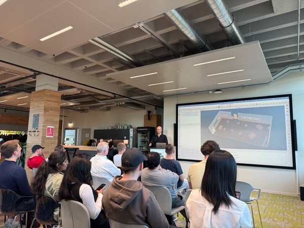 A man stands at a podium giving a presentation to a seated audience in a modern office setting with a large screen displaying a 3D model.