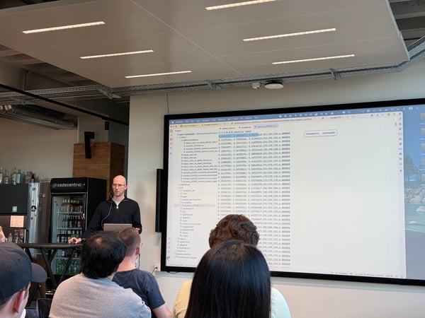 A man is giving a technical presentation in an office setting, standing next to a large screen displaying code and data to an audience.