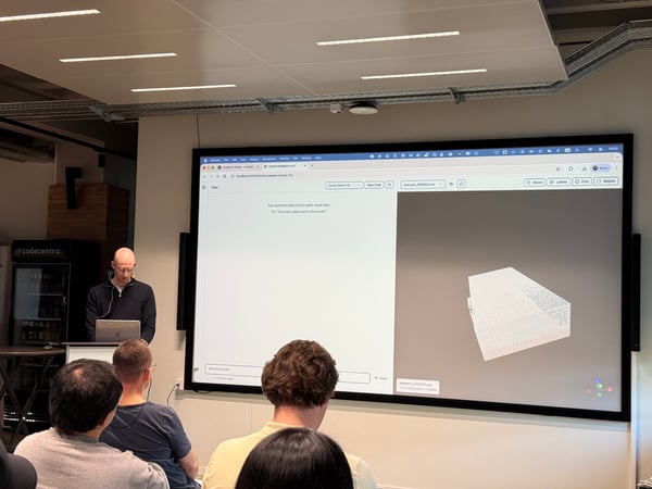 A man is giving a technical presentation or demo, standing at a podium with a laptop while a large screen behind him displays a 3D point cloud model and a chat interface. An audience is seated in the foreground, watching the presentation.
