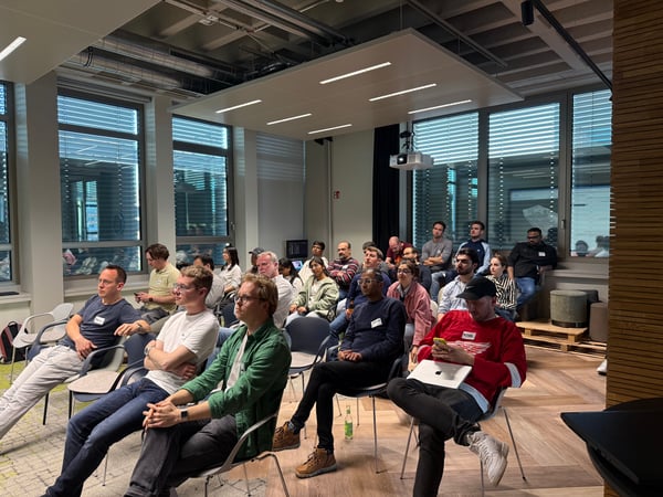 A group of people are seated in a modern office space, attentively watching a presentation or speaker off-camera.