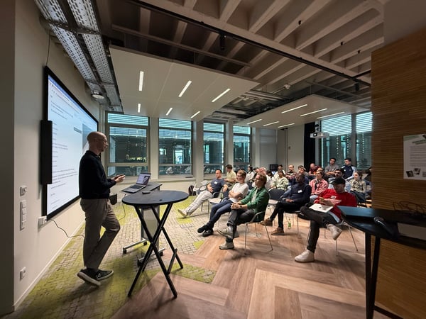 A man is giving a presentation to a seated audience in a modern office or educational space with a large screen displaying information.