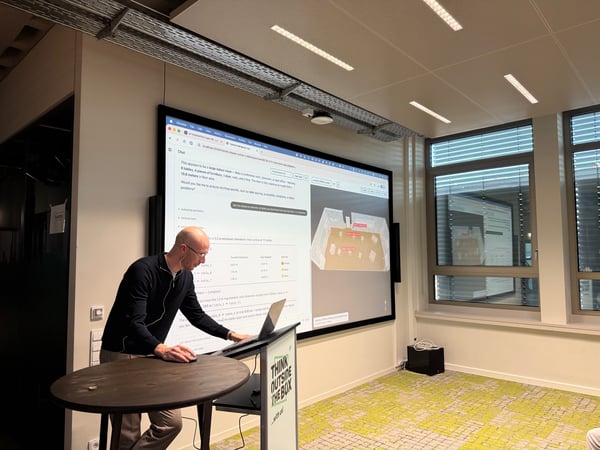 A man stands at a high table using a laptop to present a 3D room model and spatial analysis data on a large wall-mounted screen in a modern office or showroom.