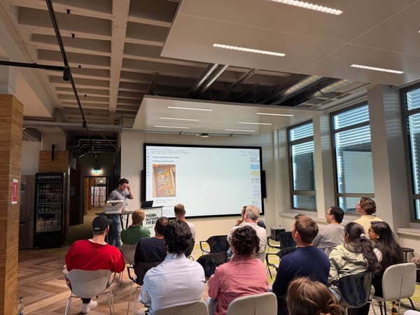 A group of people are gathered in a modern office space, watching a presentation on a large screen about selling cards online.