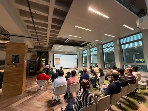 A group of people are seated in a modern office space, attending a presentation shown on a large screen.