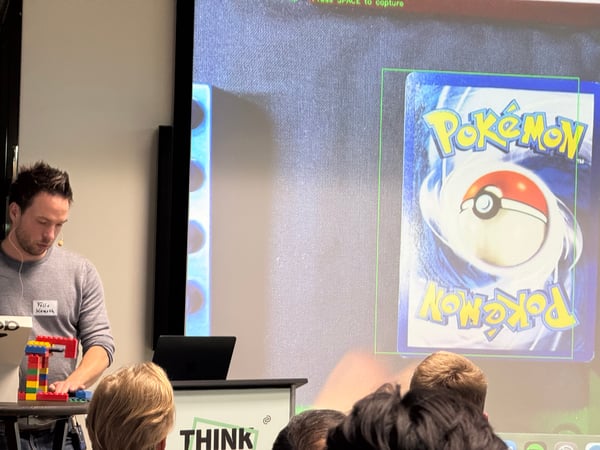 A man is giving a technical demonstration or presentation, standing next to a LEGO structure while a Pokémon card with a computer vision bounding box is projected on a screen behind him.