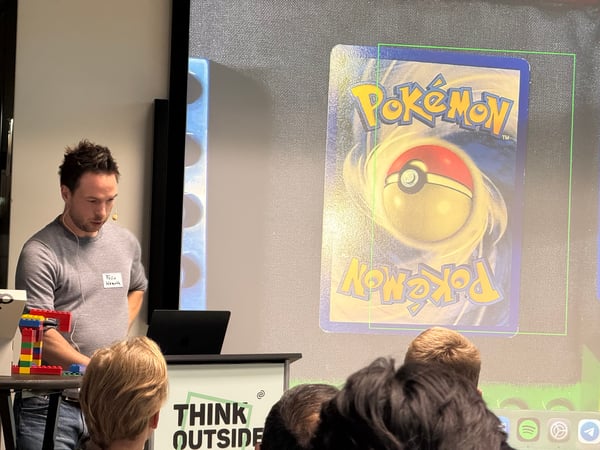 A man stands at a podium giving a presentation, with a large screen behind him displaying the back of a Pokémon trading card.