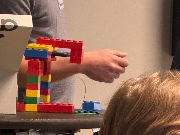 A person gestures toward a colorful LEGO structure holding a smartphone on a table during a demonstration.