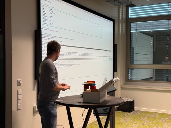 A man stands in an office setting, demonstrating a card-scanning system displayed on a large screen that shows technical logs and Pokémon card data.