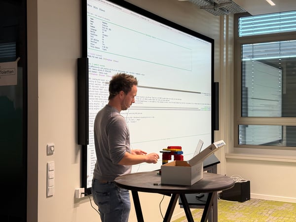 A man is standing at a high table in front of a large screen, demonstrating a technical project that appears to be a Pokémon card scanner made with LEGO bricks.